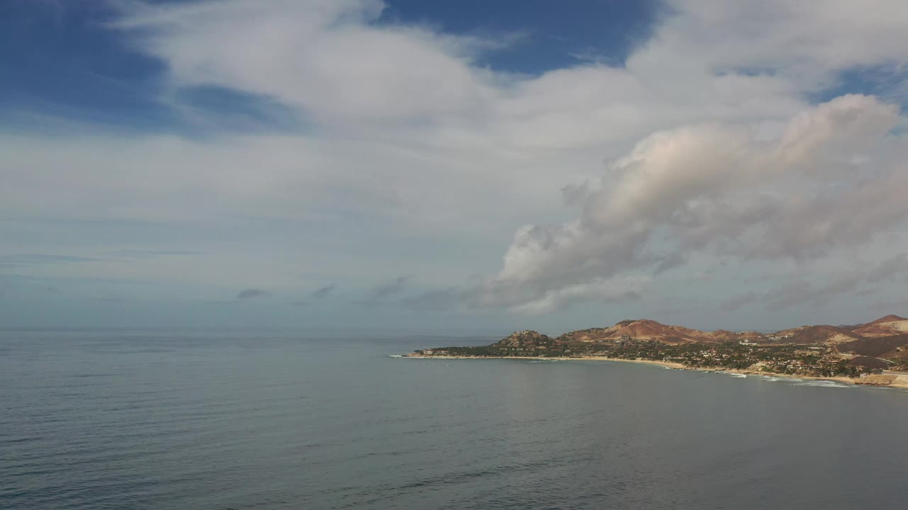 Early frame over Los Cabos, Mexico showing more open water at left, the coastline running across the lower right, brown hills beyond it, layered clouds, and no visible people.