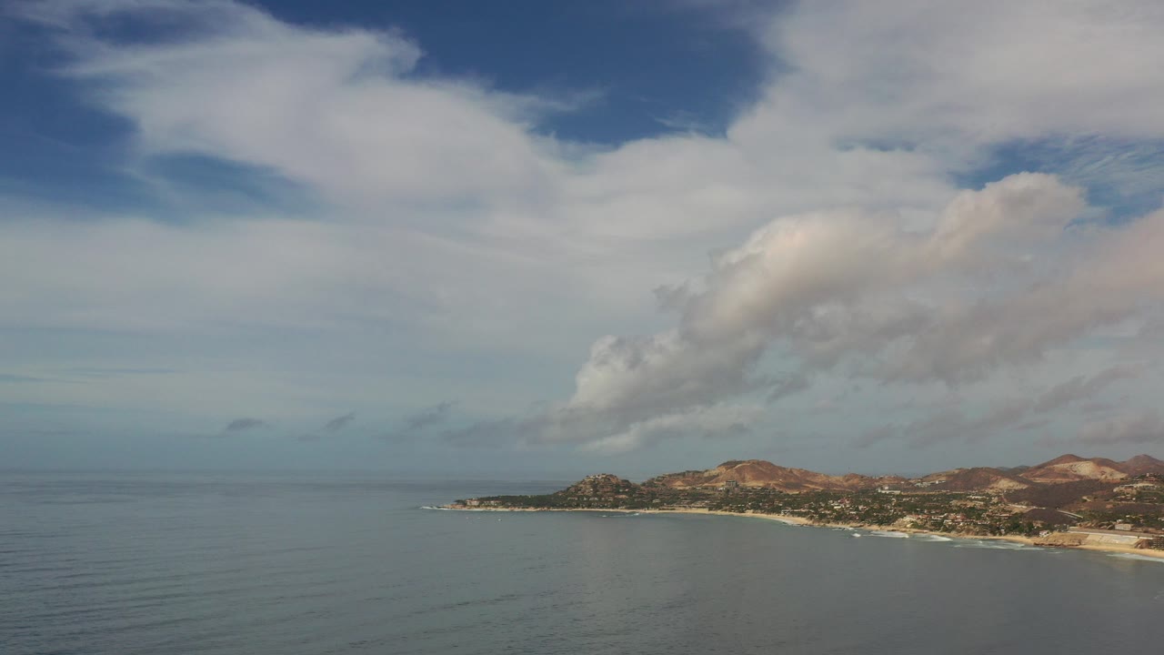 Opening aerial frame over Los Cabos, Mexico, with calm blue water in the foreground, a low coastal strip at right, reddish hills beyond, broad white clouds overhead, and no visible people.