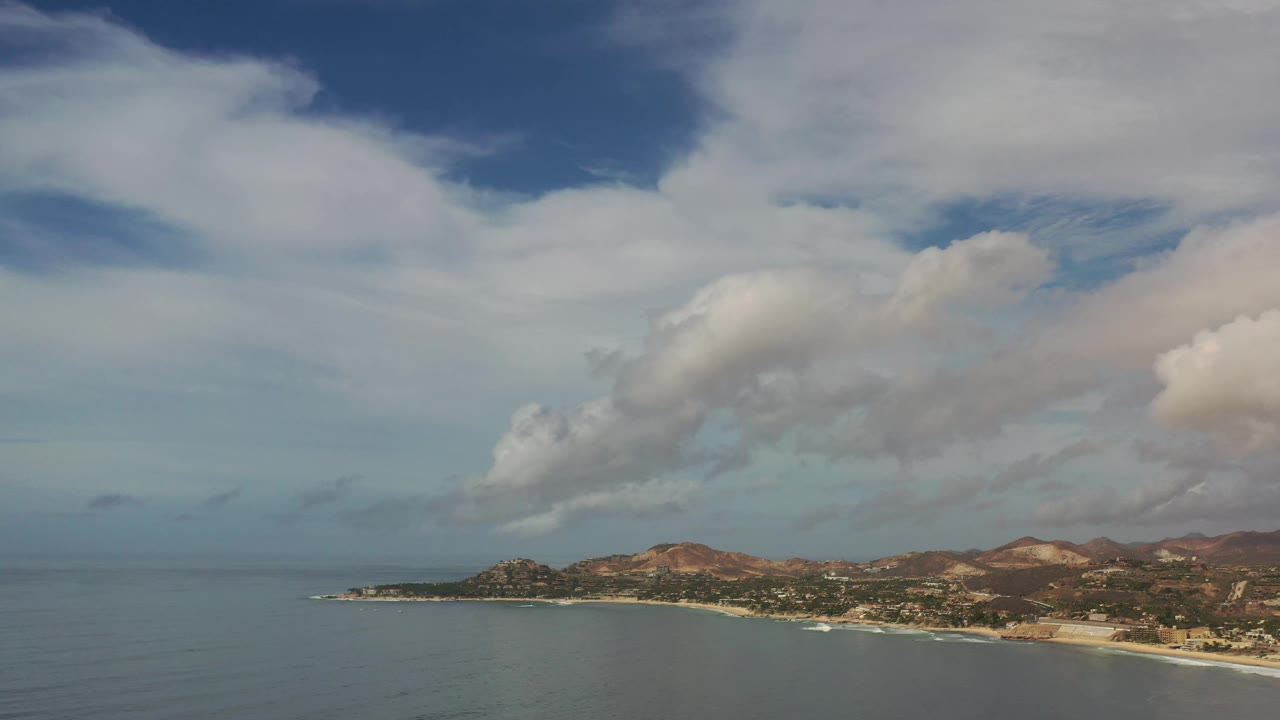 A second wide frame holds on the Los Cabos coast, showing calm blue water, a pale beach strip, low hills, distant buildings, and no visible people.