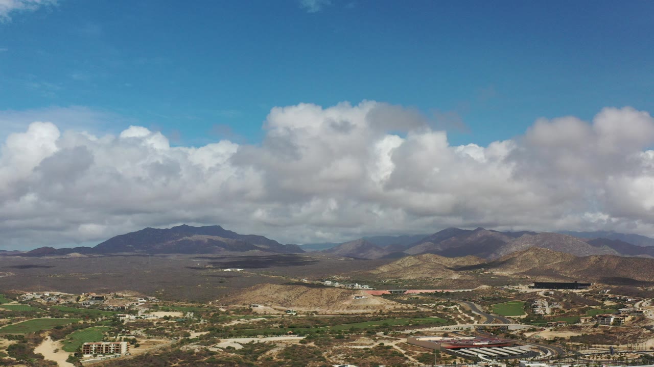 Opening aerial frame over Los Cabos, Mexico, with green planned areas and low buildings in the foreground, arid hills and mountains beyond, and no visible people.