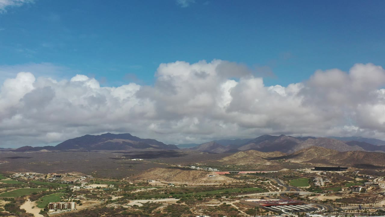 Early aerial view of Los Cabos, Mexico, showing a broad desert valley, green landscaped strips, scattered buildings, distant mountains, and no visible people.