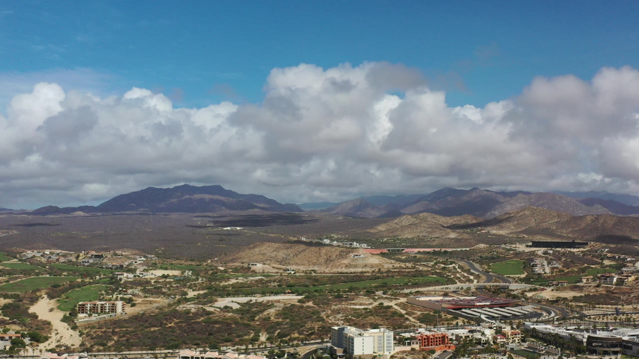 Aerial view over city buildings, desert hills, mountains, and a cloudy blue sky.
