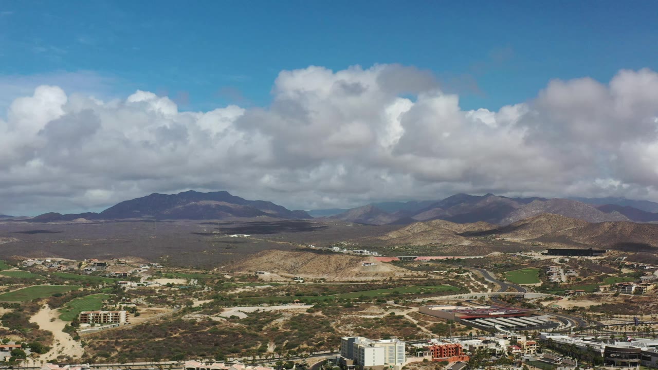 Aerial view in Los Cabos, Mexico, showing a wider band of desert terrain across the middle, built areas near the lower edge, mountains on the horizon, and no visible people.