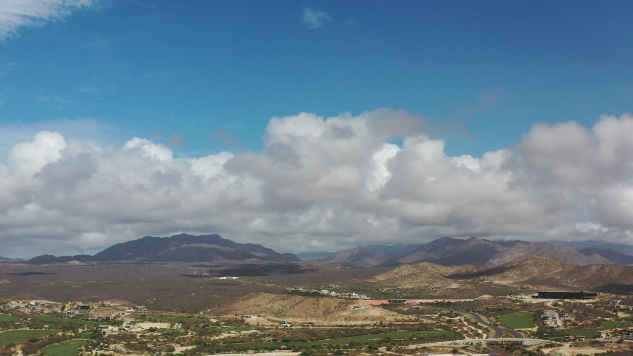 Opening frame of a Los Cabos, Mexico mountain panorama with dry hills, green fields, pale roads, scattered buildings, blue sky, large clouds, and no visible people.