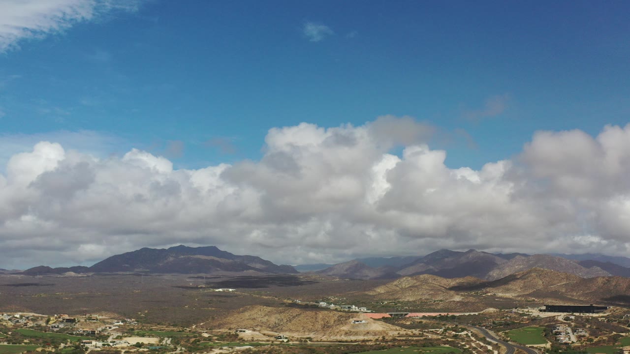Wide aerial view of dry hills, roads, distant mountains, and large clouds.