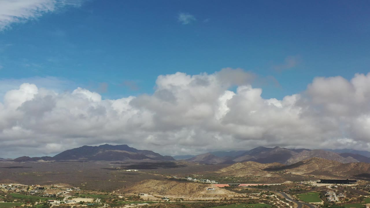 Los Cabos aerial frame with the central mountain ridge slightly more prominent, cloud shadows across dry hills, scattered buildings, roads, and no visible people.