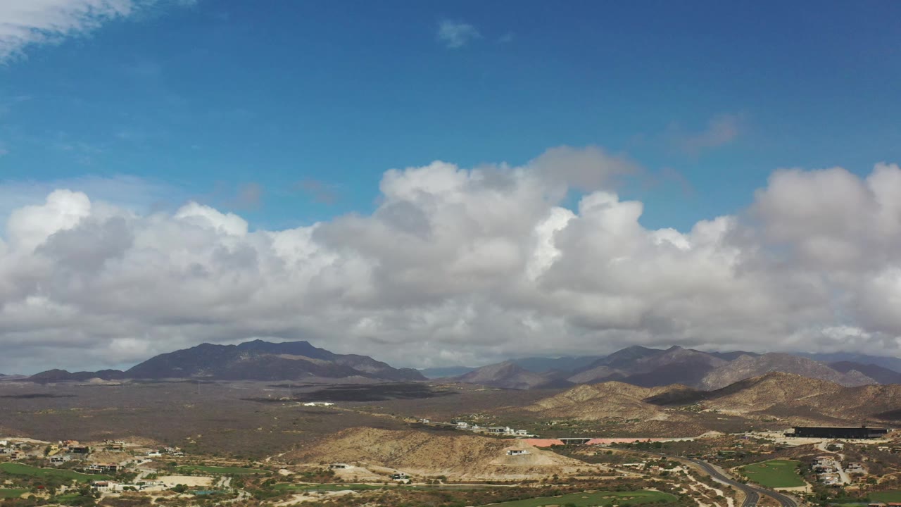 Elevated Los Cabos landscape with large white clouds crossing the blue sky, brown mountain ridges, light roads, sparse buildings, and no visible people.