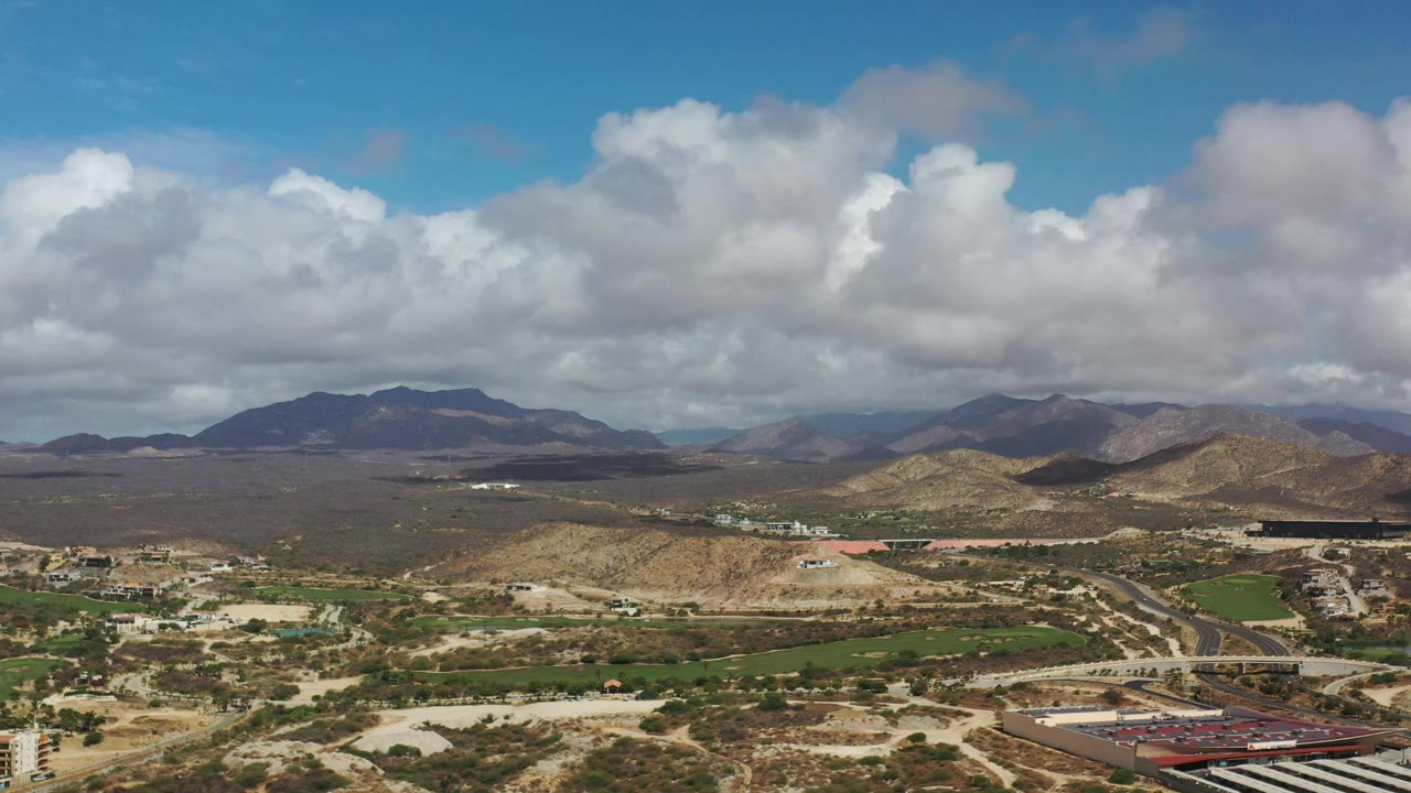 Wide drone view in Los Cabos, Mexico, with tan ridge lines across the foreground, green agricultural-looking plots below, mountains on the horizon, and no visible people.