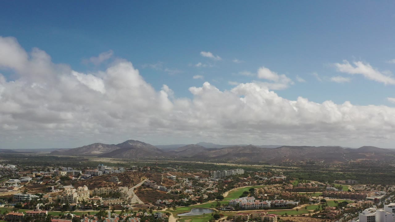 Wide aerial view of city buildings, green space, mountains, and cloud-filled sky.