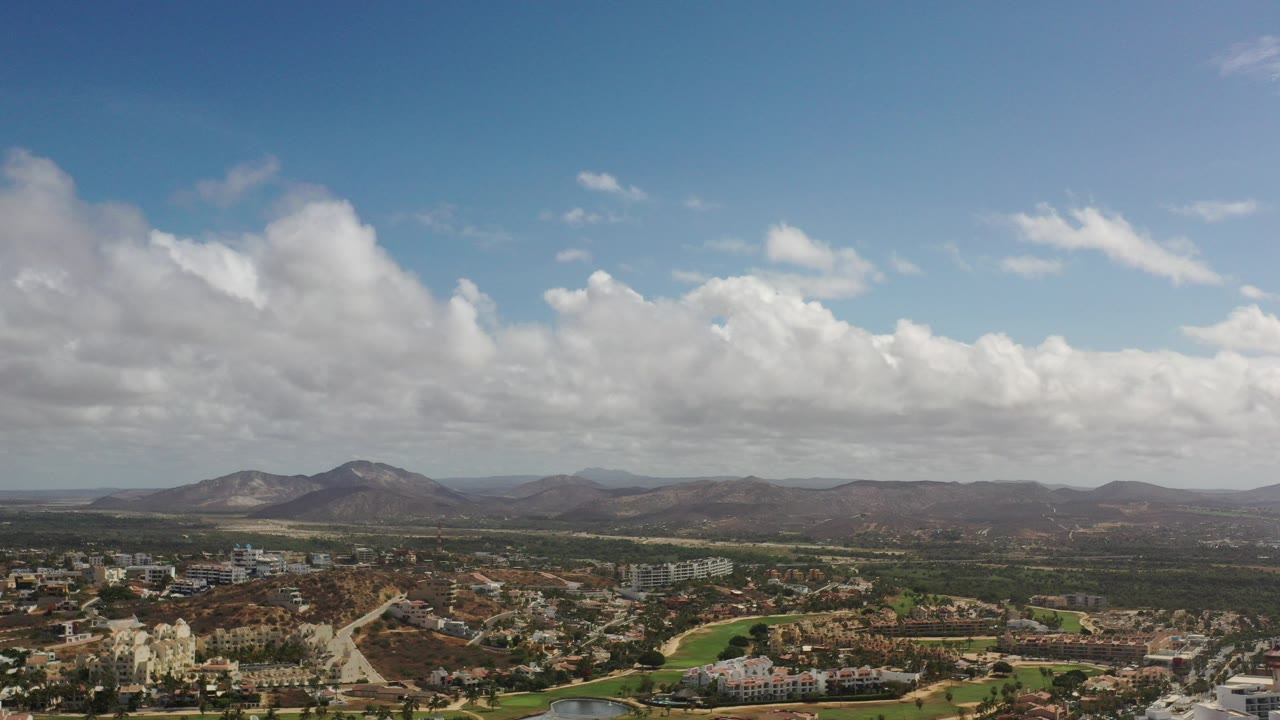 Opening aerial frame over Los Cabos, Mexico, with low-rise buildings and roads below, dry hills and mountains on the horizon, a blue sky with large white clouds, and no visible people.
