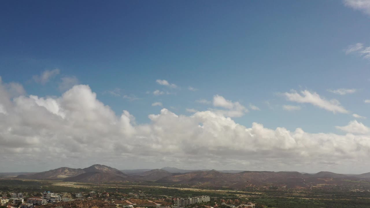 Los Cabos mountain view with a broad bank of white clouds, muted brown hills, a thin built-up strip near the bottom, and no visible people.