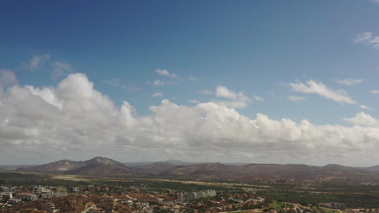 Opening aerial frame over Los Cabos, Mexico, with low buildings at the bottom edge, green fields across the midground, distant hills, large clouds, and no visible people.
