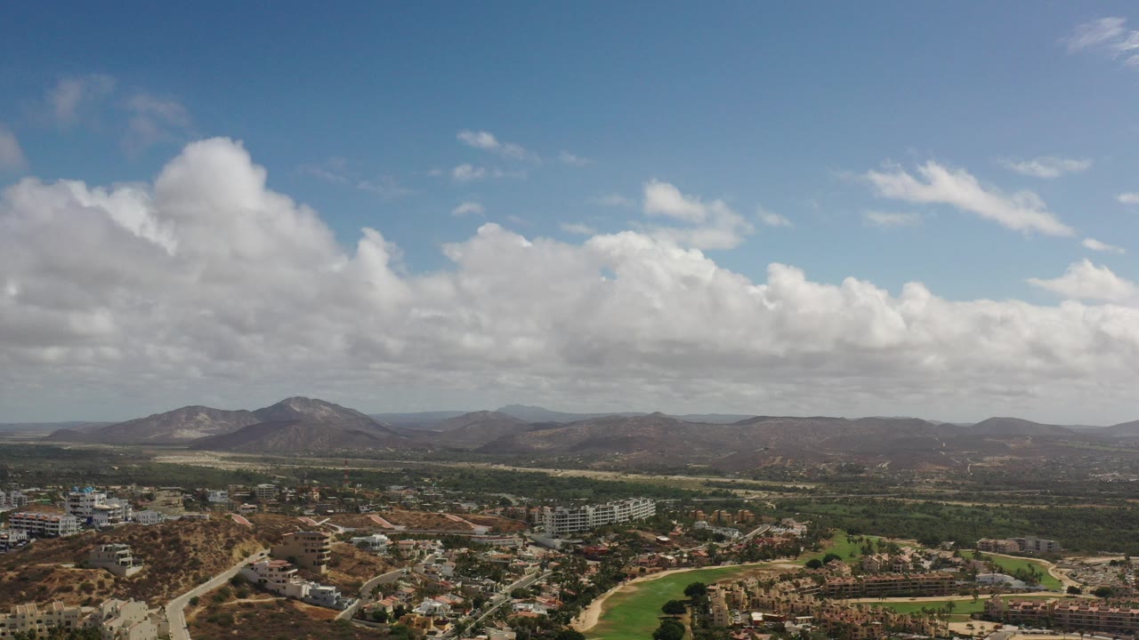 Aerial view of green grass, buildings, hills, mountains, and a cloud-filled sky.