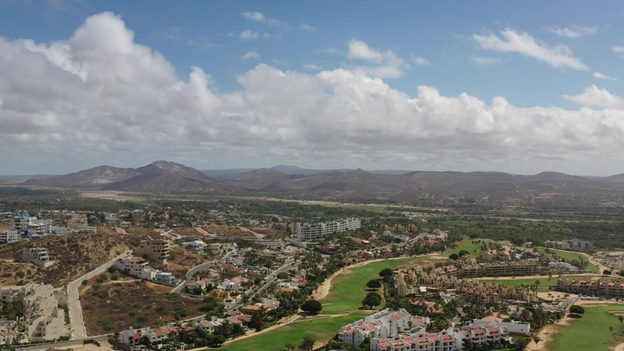 Opening frame of Los Cabos from above, showing roads, low buildings, green landscaped strips, dry hills, distant mountains, large clouds, and no visible people.
