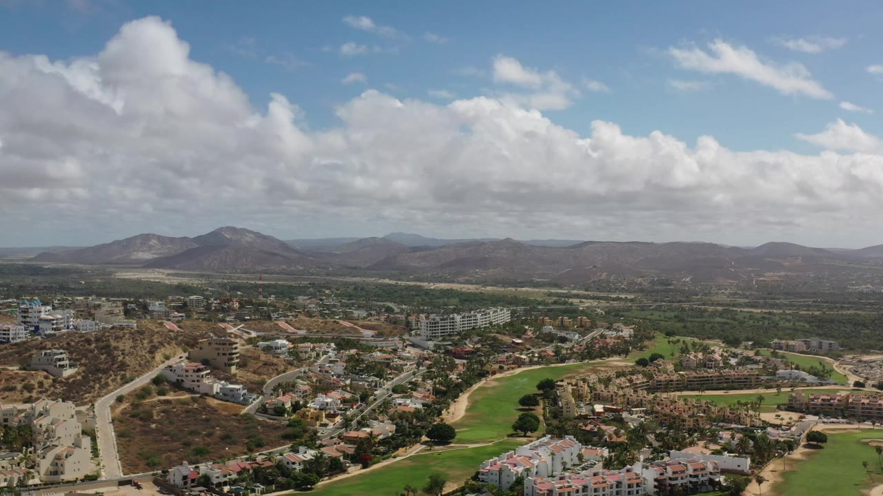 Los Cabos aerial frame with a central road descending through buildings, bright green fairway-like grass, brown hills, mountains on the horizon, and no visible people.