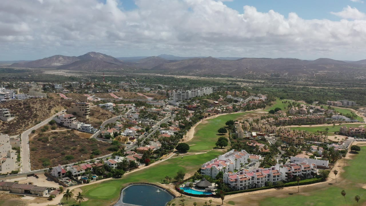 Aerial view of homes, golf greens, pond, roads, dry hills, and mountains.