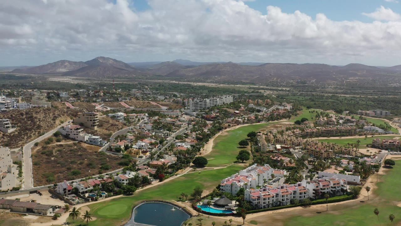 Aerial frame over Los Cabos with the pond now nearer the lower center, white residential blocks in front of fairways, curving roads, dry hillsides, mountains, and no visible people.