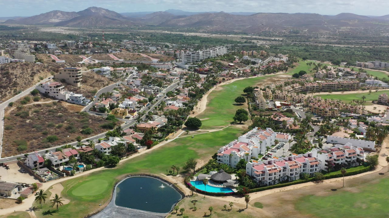 Aerial view over Los Cabos, Mexico, showing a golf green, dark pond, white apartment buildings with pink roofs, hillside roads, distant mountains, and no visible people.