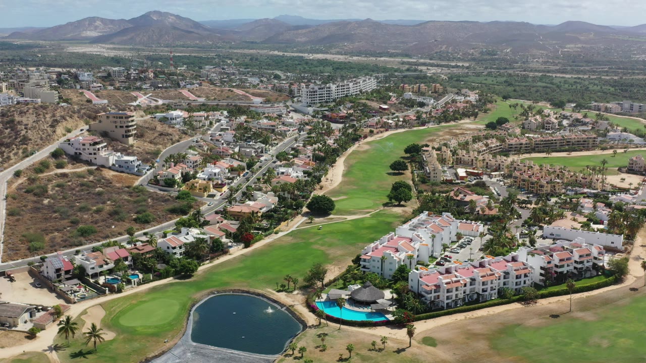 Aerial view in Los Cabos, Mexico, with the pond centered low in frame, turquoise pool beside white apartment blocks, green fairway, neighborhood roads, dry hills, and no visible people.
