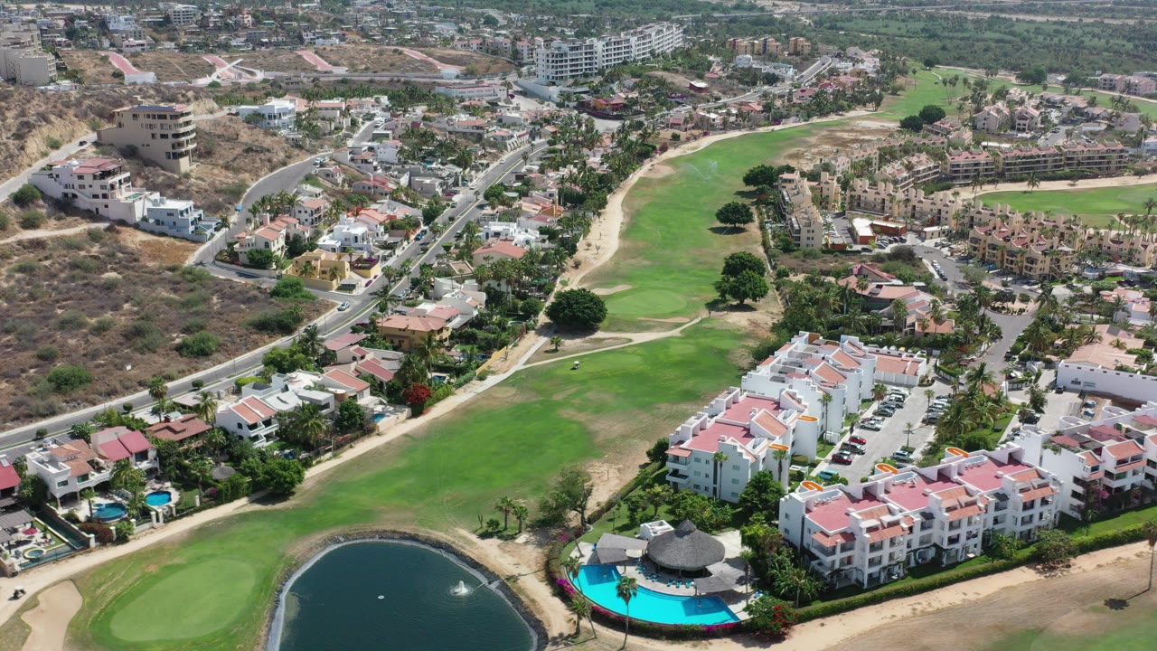 Opening high aerial frame over Los Cabos golf fairways with a pond at the lower edge, red-roof resort buildings, roads, hillside homes, and no visible people.