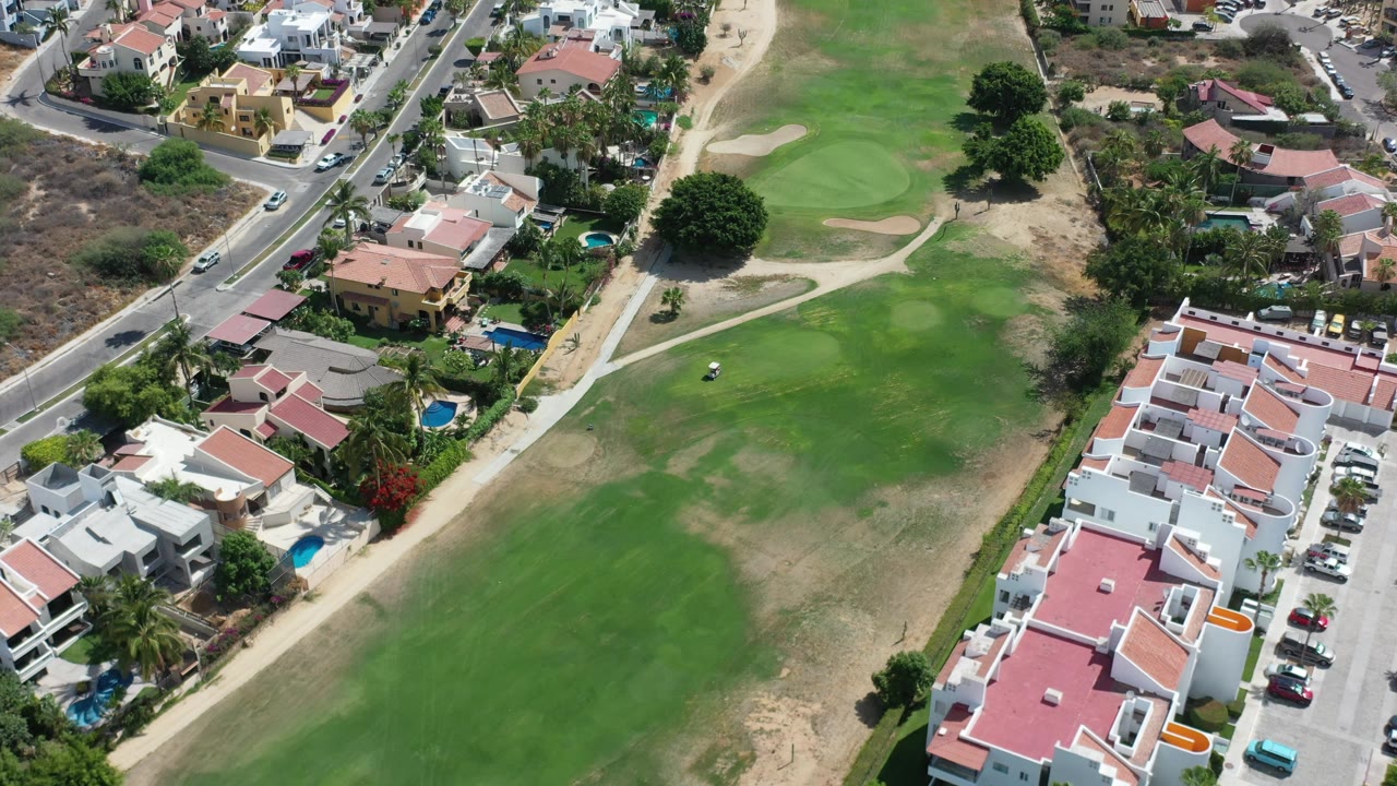 Aerial frame in Los Cabos, Mexico, with the green field filling most of the view, houses along the right side, a road near the top, and no visible people.