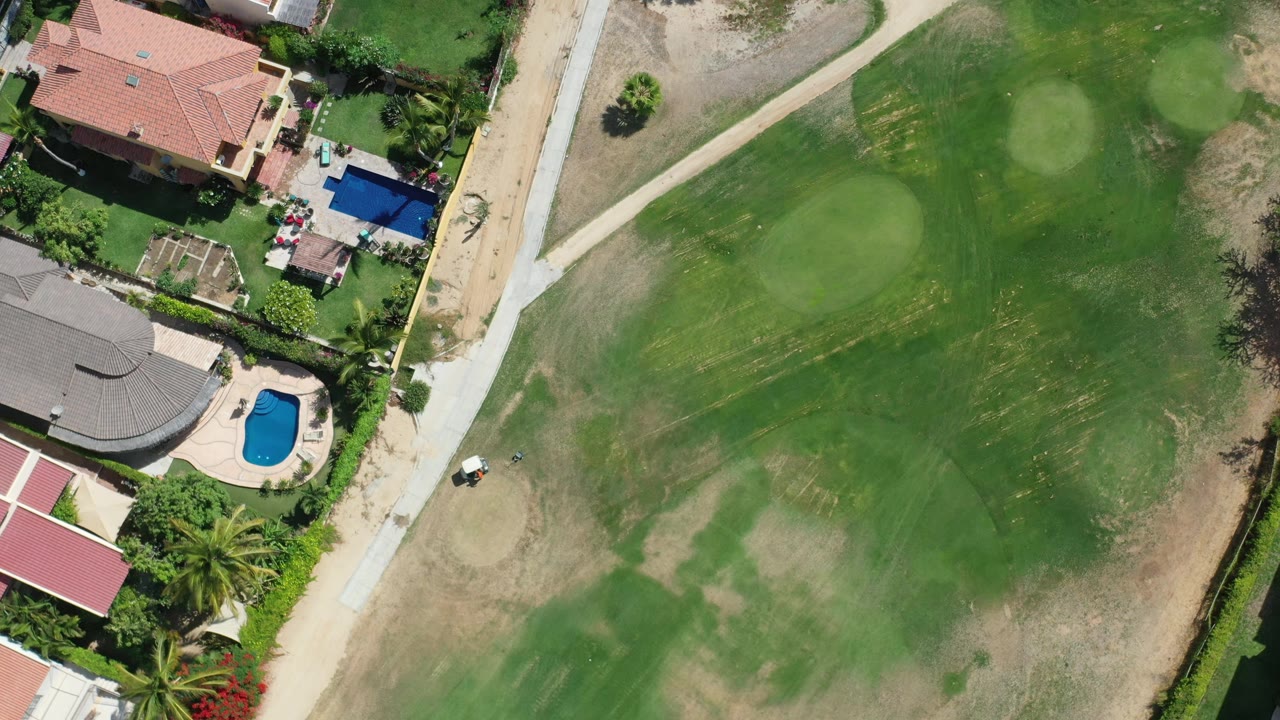Aerial frame over Los Cabos, Mexico with a putting green and fairway next to red-roof and gray-roof homes, blue pools, paths, and no visible people.