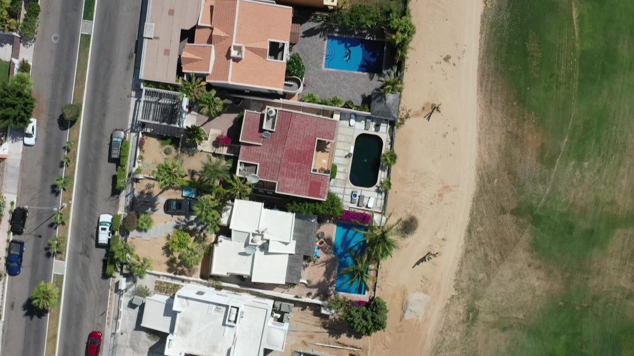 Overhead daytime view in Los Cabos, Mexico, showing private homes with pools beside a paved road and a broad green strip, with no visible people.