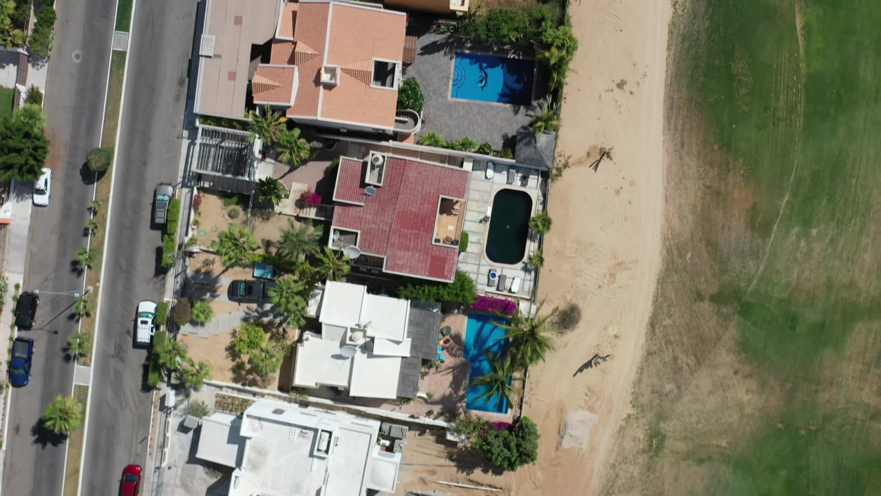 Top-down Los Cabos residential frame with tan and red roofs, blue pools, palm landscaping, parked vehicles, road pavement, and no visible people.