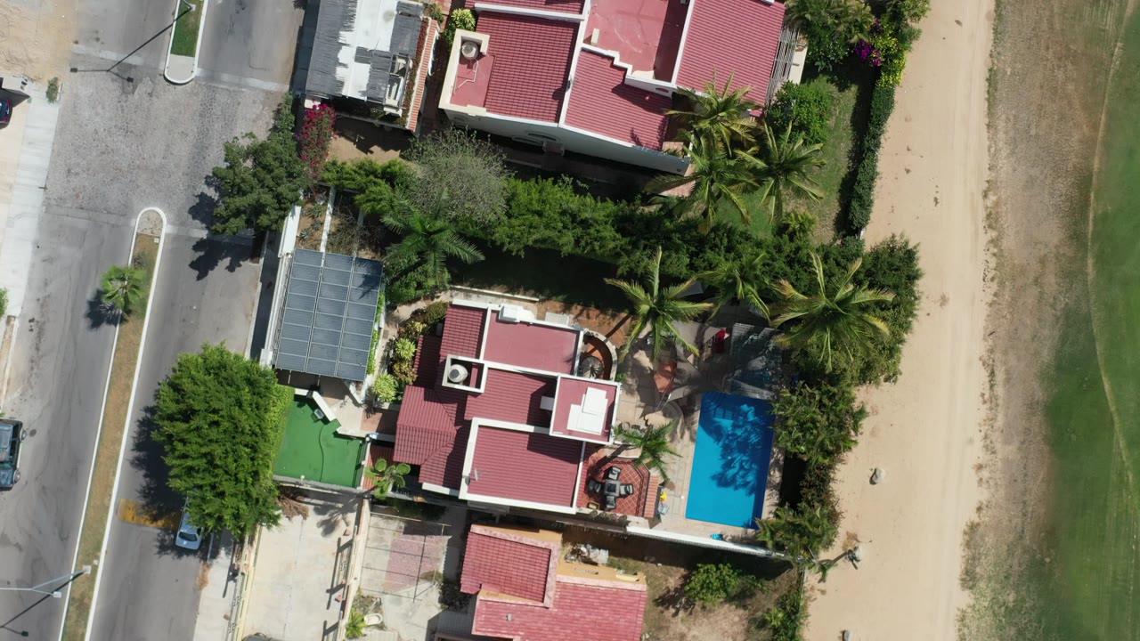 Aerial frame in Los Cabos, Mexico showing the red-roofed house and pool slightly farther along the overhead view, bordered by palms, a road, and nearby buildings, with no visible people.