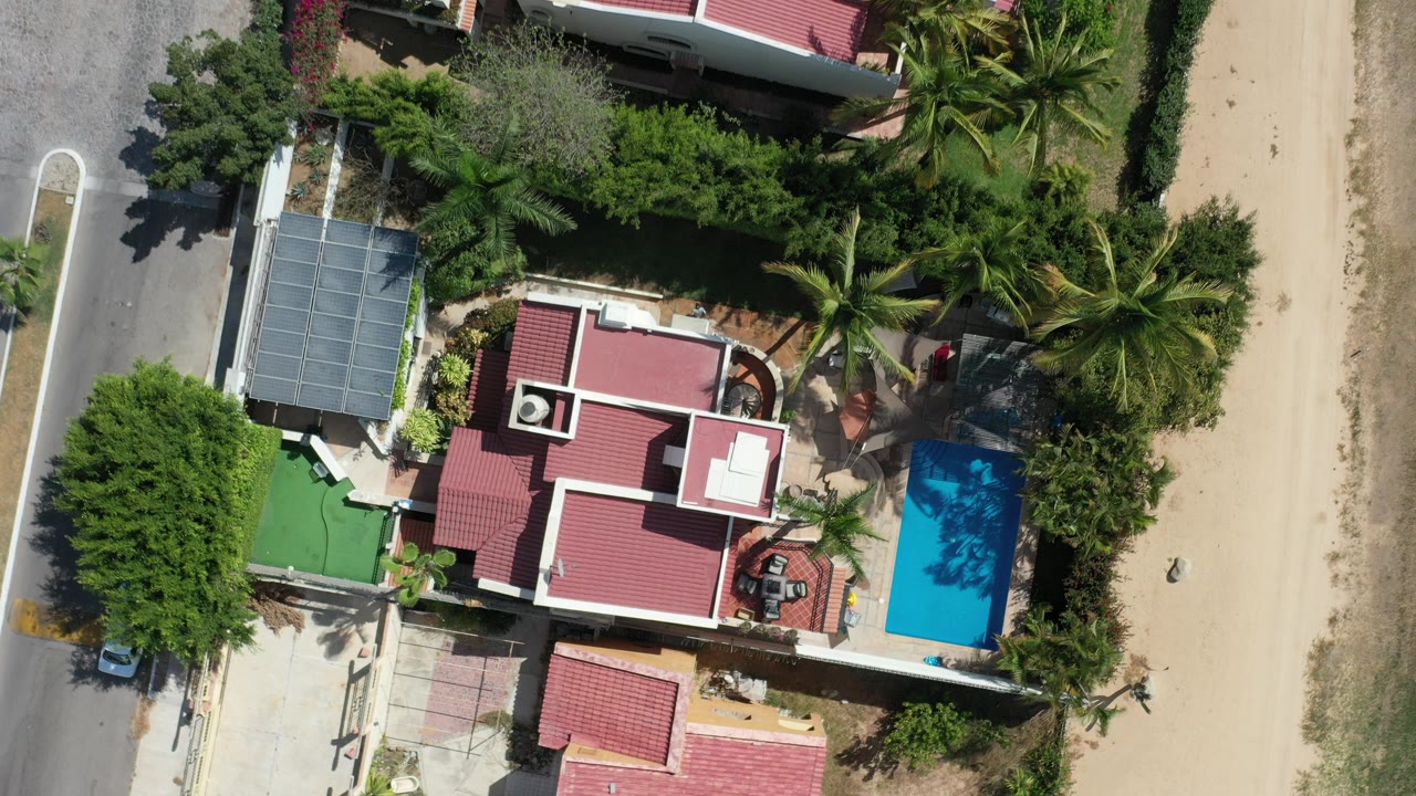 Overhead frame of a Los Cabos beachside home with red roofs, a blue pool, palm trees, a paved road, sandy beach, and no visible people at the start of the clip.