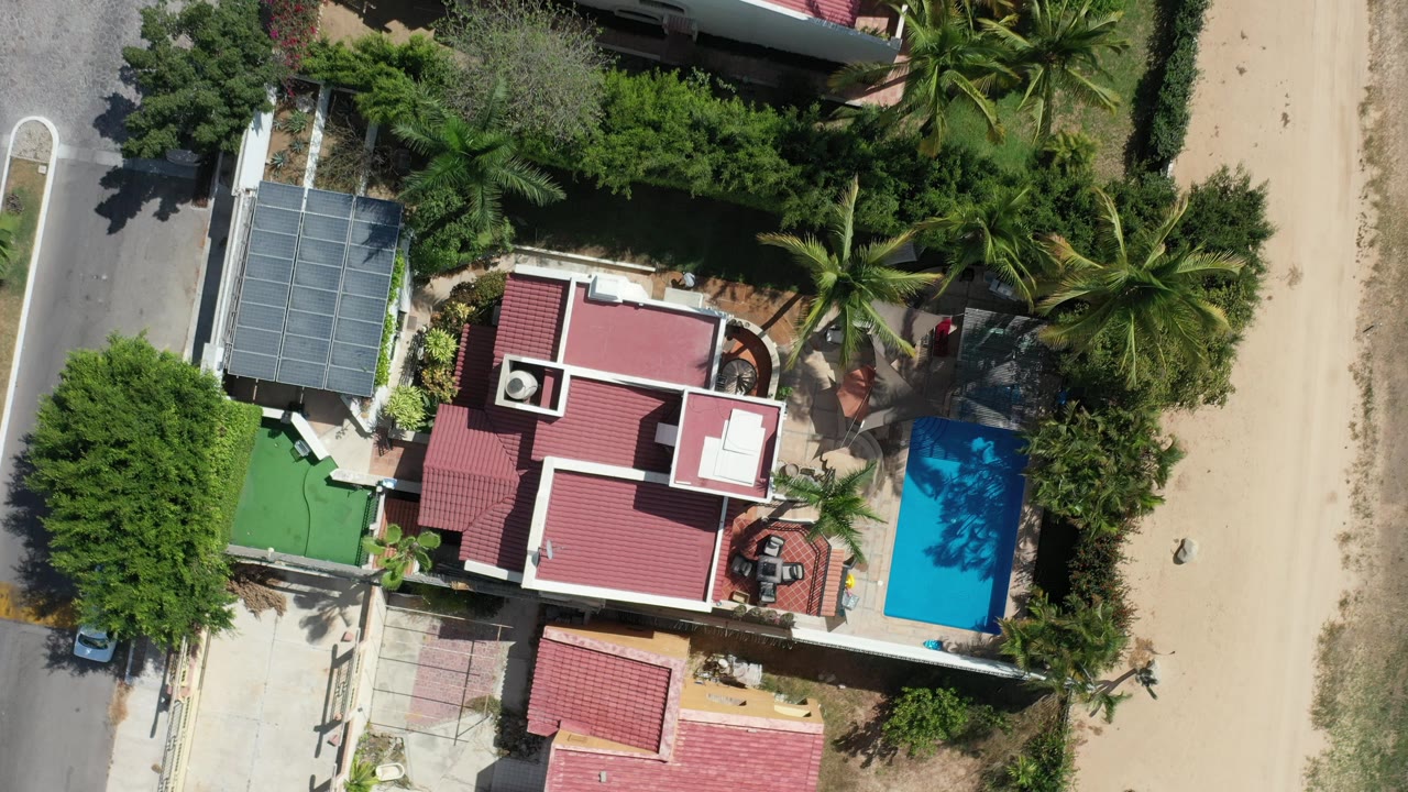 Top-down shot of a beachside house, blue pool, palms, road, and sand.
