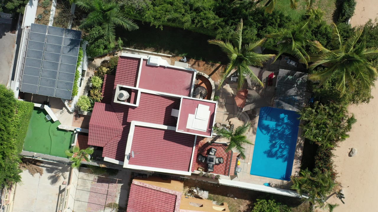 Overhead aerial frame in Los Cabos, Mexico showing a red-roofed house, blue rectangular pool, palms, pale paths, and no visible people.
