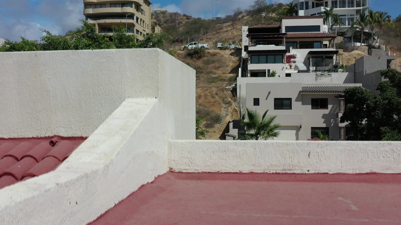 Red roof and white stucco parapet with hillside homes behind it.