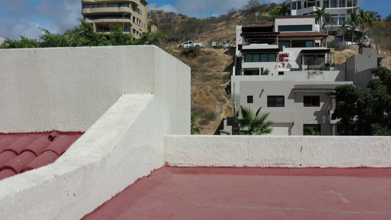 At 0:01, the white stucco wall cuts across the red rooftop while modern homes on the Los Cabos hillside remain visible beyond it, with no visible people.