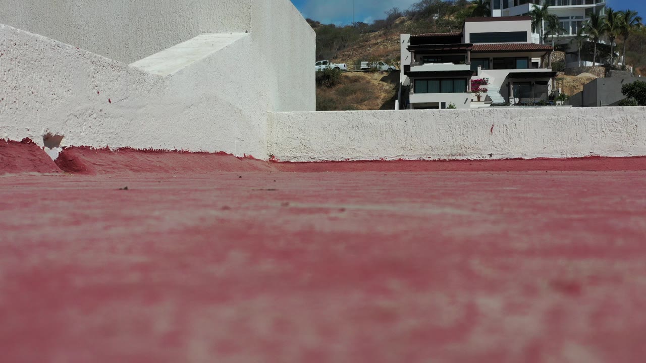 Low red pavement and white wall with hillside homes behind it.