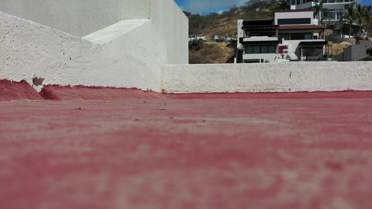 Los Cabos terrace frame with the red floor dominating the lower half, a rough white parapet across the center, background homes on a dry slope, and no visible people.