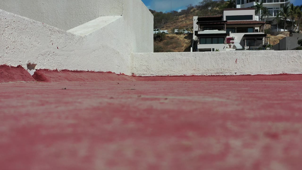 A near-static Los Cabos rooftop frame keeps the red foreground soft, the white parapet sharp across the middle, and modern homes on the dry hillside with no visible people.