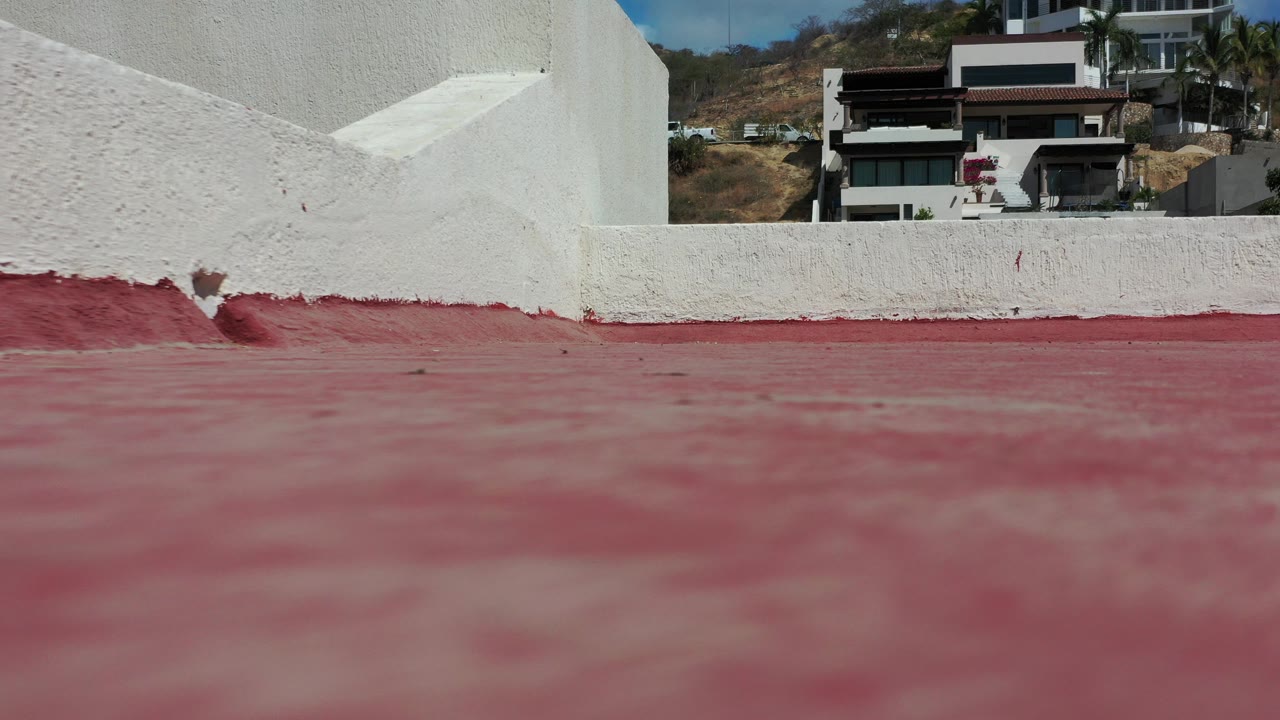 Low rooftop view in Los Cabos, Mexico, showing a red roof surface, white stucco parapet, hillside homes, blue sky, and no visible people.