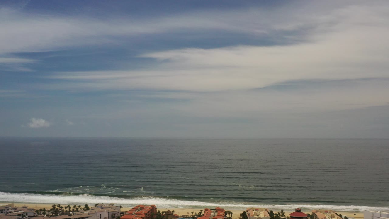 Wide elevated view over the Los Cabos coastline, with blue-gray ocean, a thin beach edge, rooflines at the bottom, broad clouds above, and no visible people.