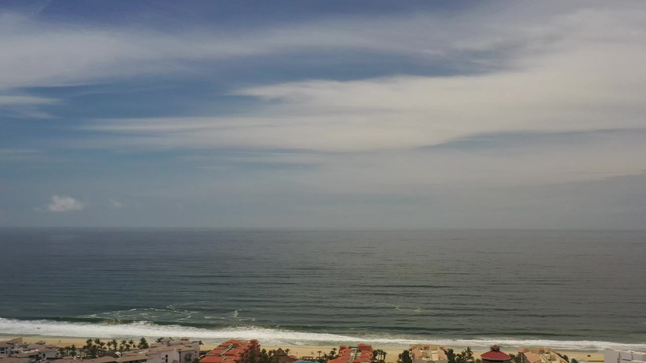 Elevated Los Cabos coast view with the ocean stretching to a hazy horizon, white surf near the beach, small palm shapes and low rooflines at the lower edge, and no visible people.