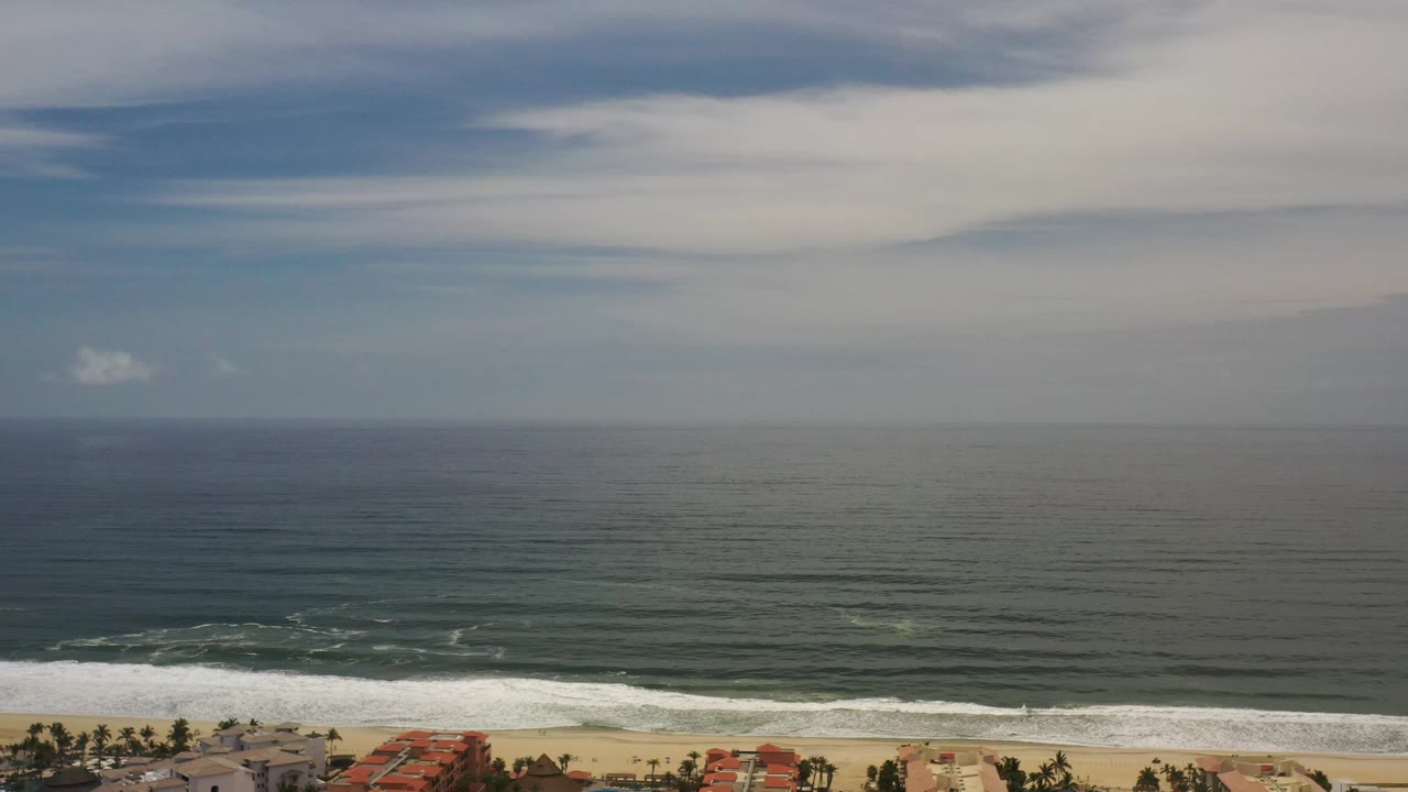 One second into the Los Cabos clip, broad cloud bands sit over calm offshore water while white waves break along the beach, with no visible people.