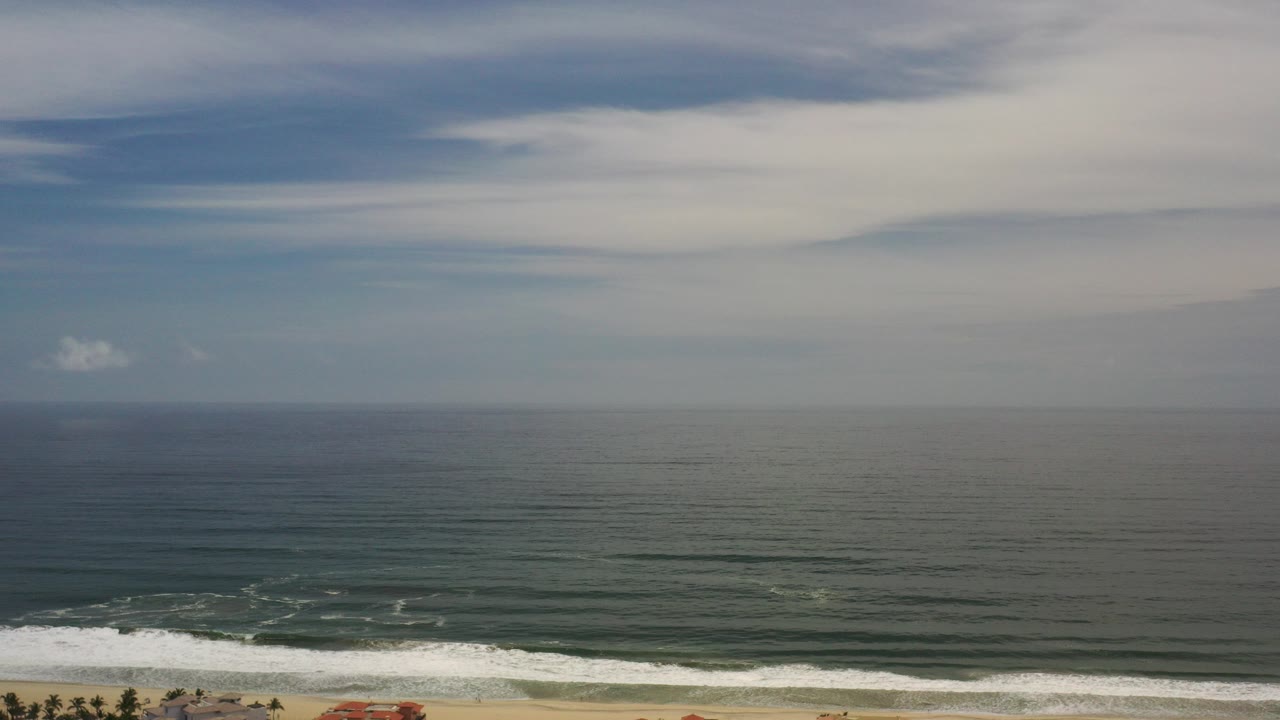 Opening Los Cabos frame with a wide blue-green ocean horizon, pale layered clouds across the sky, a thin beach and surf line at the bottom, and no visible people.