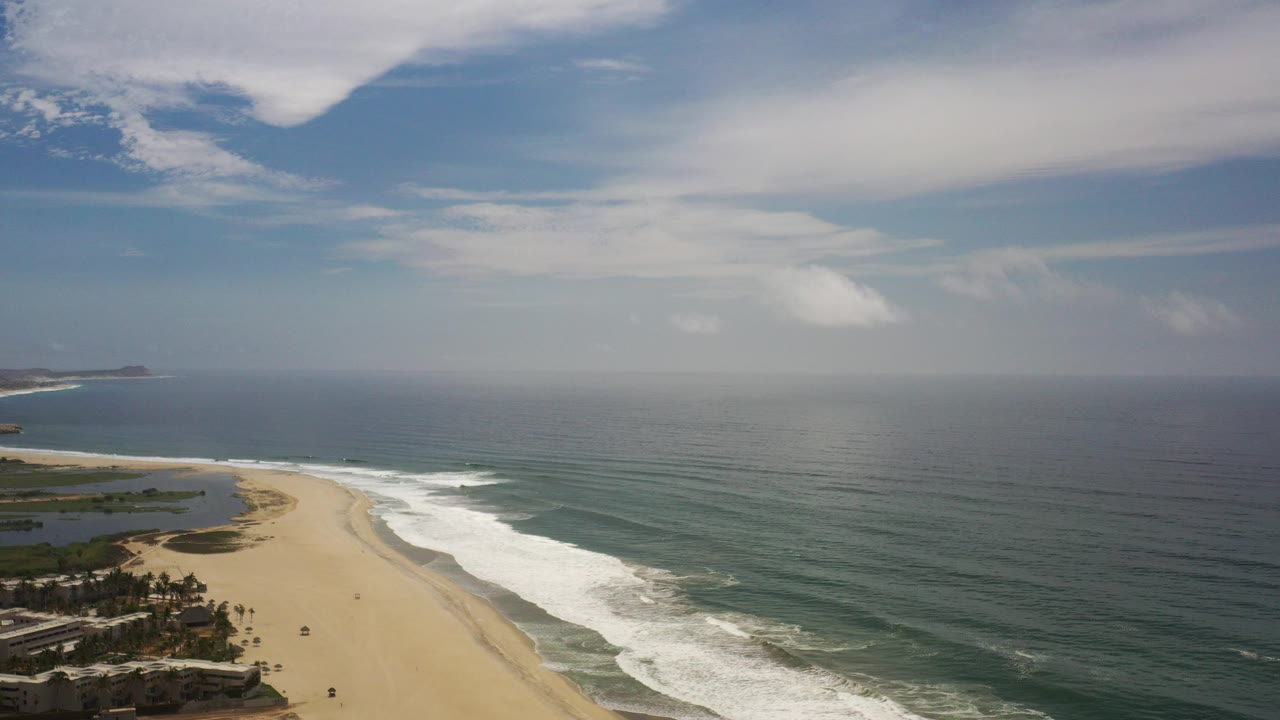 Opening aerial view over the Los Cabos coast, with sandy beach at lower left, white surf lines, open green-blue ocean, distant headland, clouded sky, and no visible people.