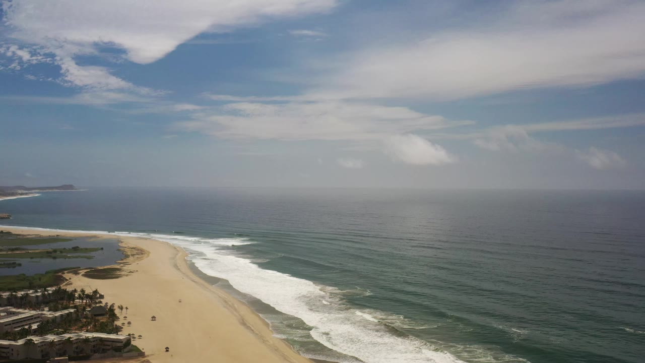 A wide Los Cabos aerial frame shows the beach curving from the lower left, waves breaking along shore, open ocean across the right side, layered clouds above, and no visible people.