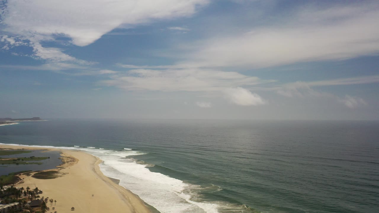 Aerial view over a Los Cabos beach with pale sand at lower left, white surf along the shoreline, open ocean to the right, a distant headland, and no visible people.