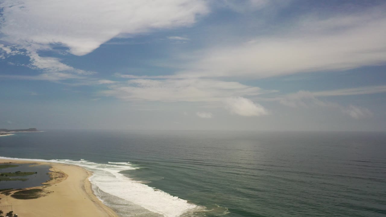 Elevated coastal view with sandy beach, white surf, ocean, distant land, and cloudy sky.