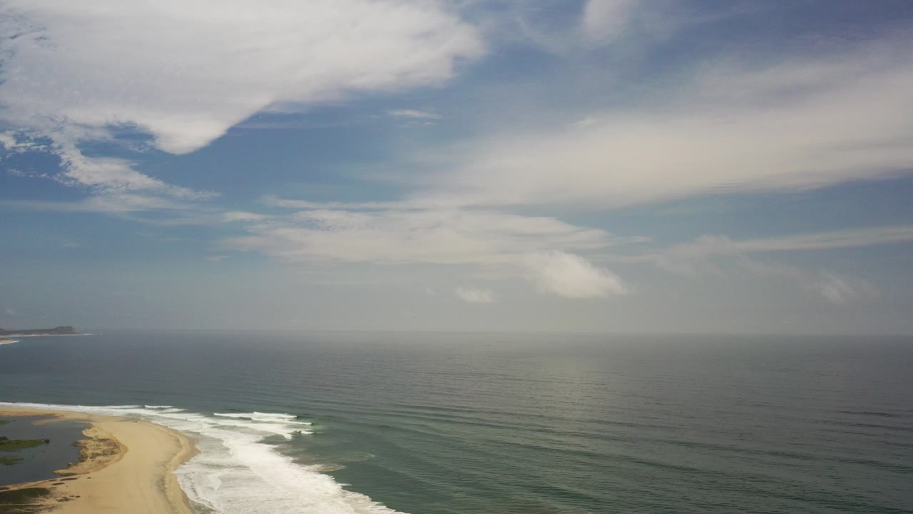Aerial coastline with sandy beach, surf, ocean, horizon, and clouds.