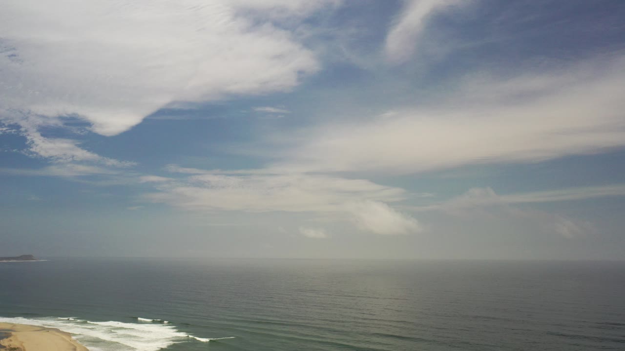 Wide ocean horizon with clouds above and surf at a sandy beach edge.