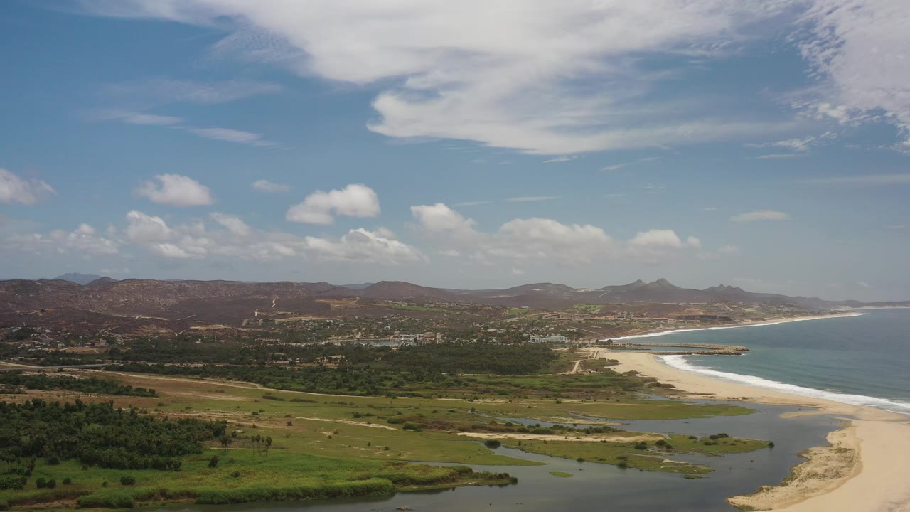 Wide aerial view in Los Cabos, Mexico, showing a sandy beach curving beside blue water, a dark lagoon in front, green plains, low hills, broken clouds, and no visible people.