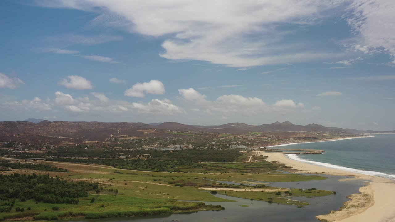Los Cabos aerial frame with the lagoon and sandbar across the lower frame, green vegetation inland, a pale beach along the water, distant brown hills, and no visible people.